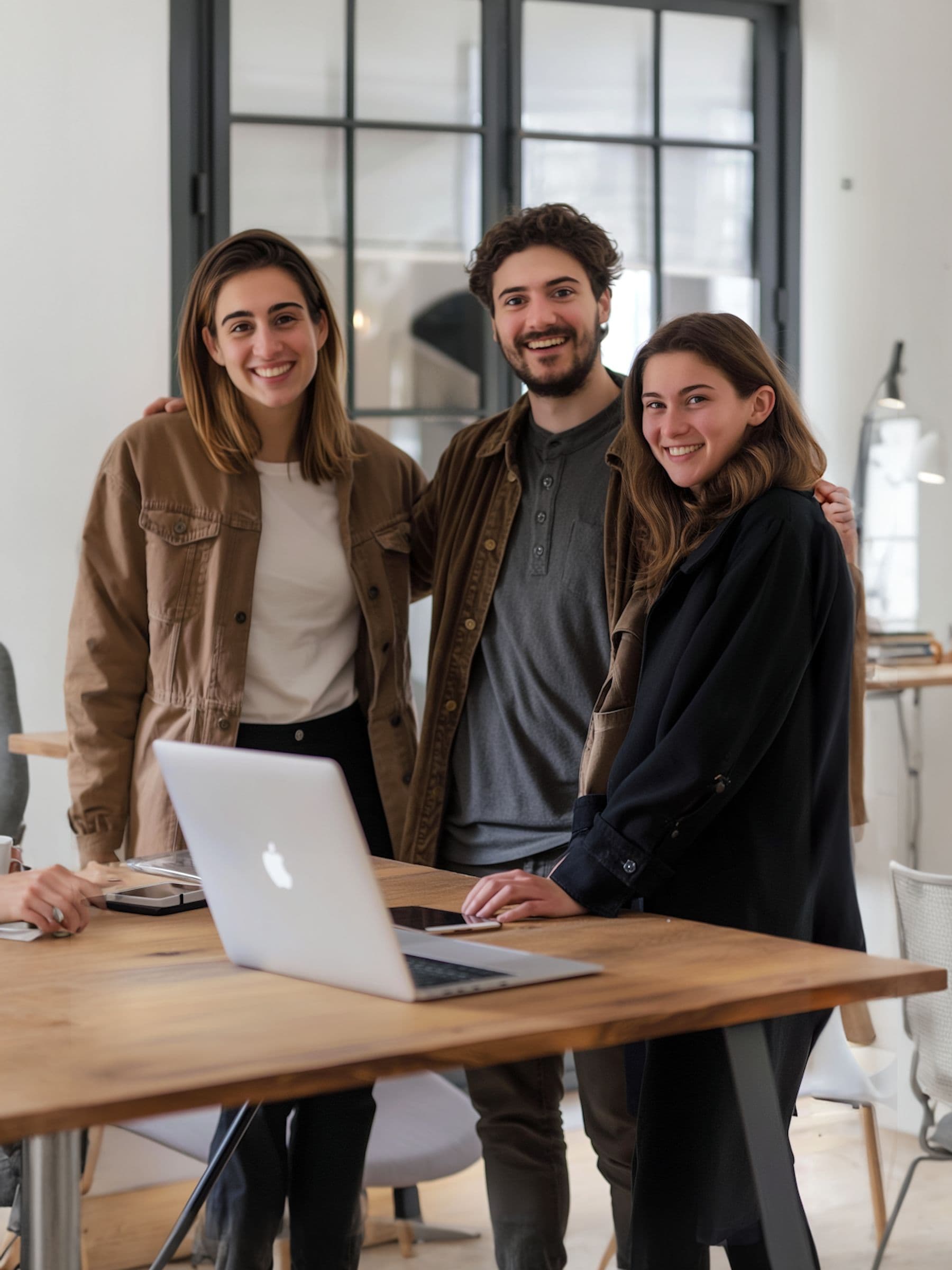 Three people smiling, standing around a laptop on a wooden table in a modern office with large windows in the background.